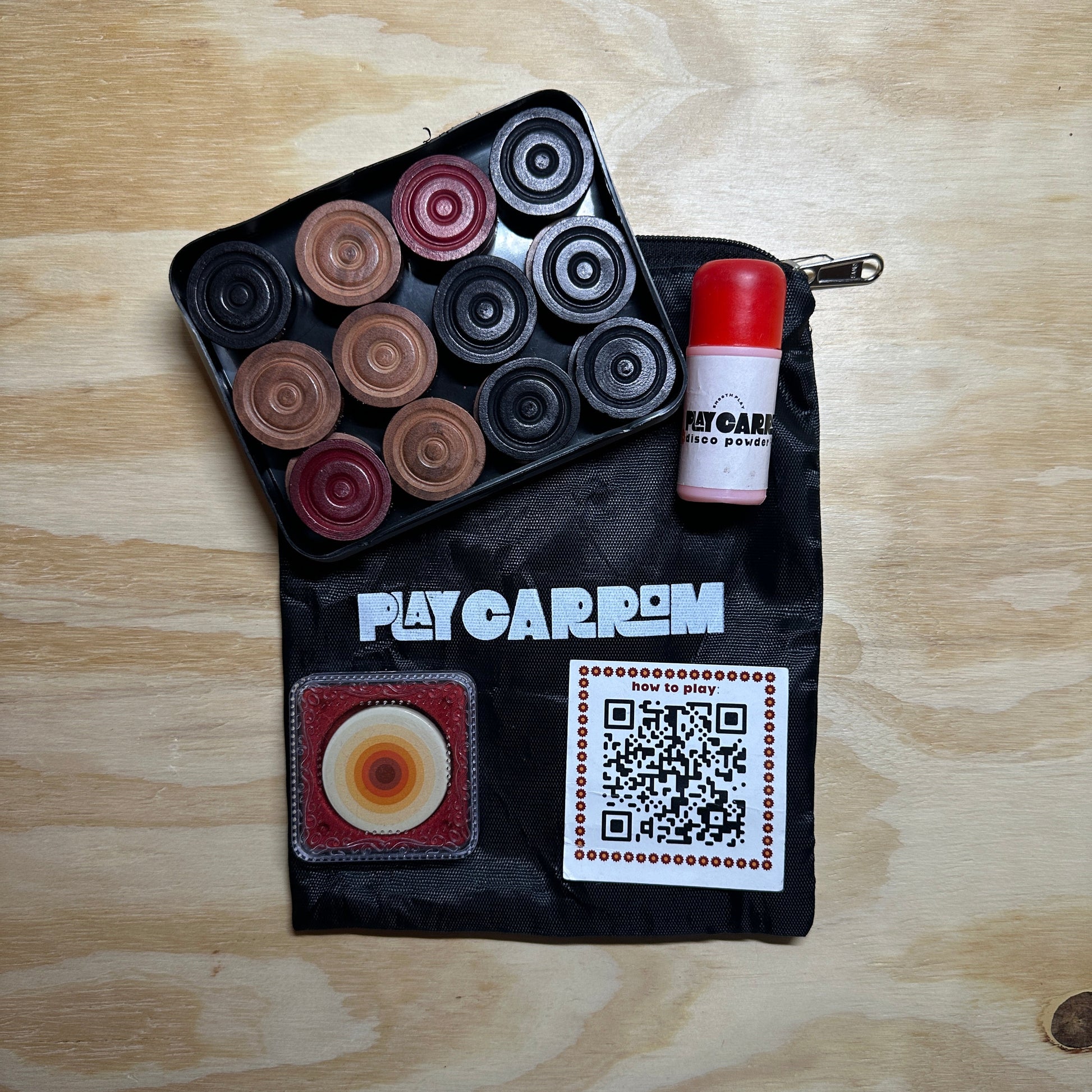 Black pouch with colorful circular carrom men, striker, and a QR code on a wooden surface