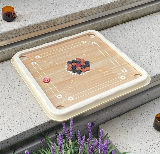 A carrom board with a wrap-around coin holder, placed on a stone surface with purple flowers in the foreground.